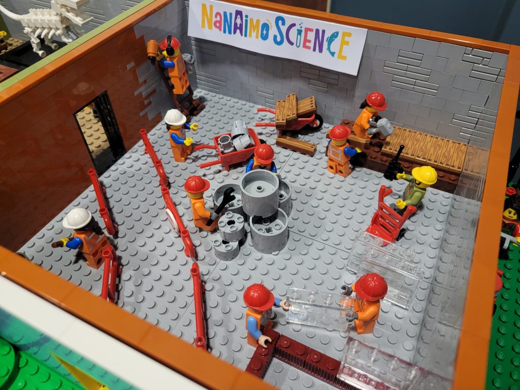 Exhibit under construction at a LEGO built Science Centre with minifigures in hard hats busy painting, hammering, and installing the exhibit.