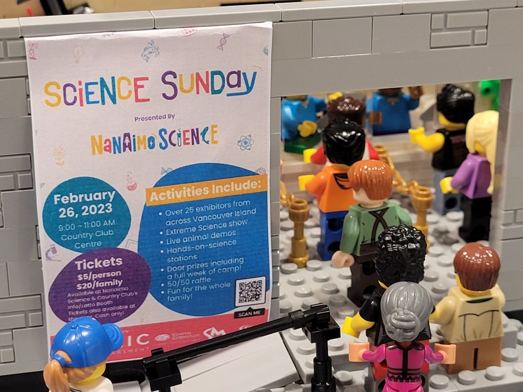 LEGO-sized Science Sunday poster on the front of the minifigure scale LEGO Science Centre with minifigures lining up through the door.